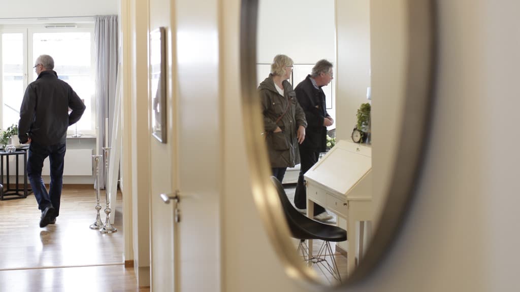 Två människor med ytterkläder syns i spegeln i en hall. En man syns bakifrån.