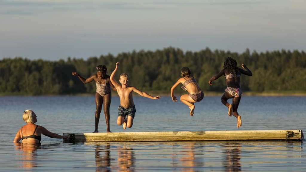 Barn med mörkbrun och ljus hy hoppar i vattnet från brygga i sjö i kvällsljus. En vuxen i vattnet.