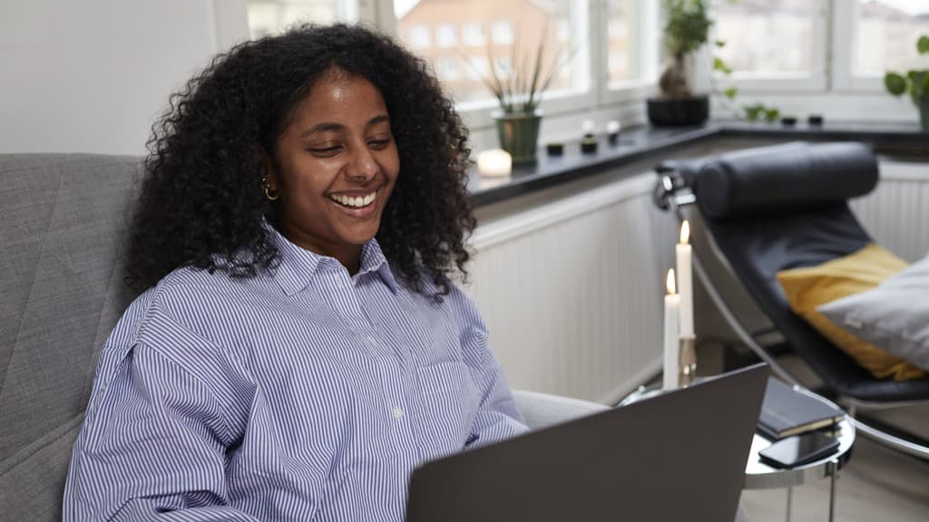 Kvinna med brun hy och vågigt mörkbrunt hår som sitter i en soffa i en lägenhet med laptop i knät och ler stort medan hon tittar på skärmen