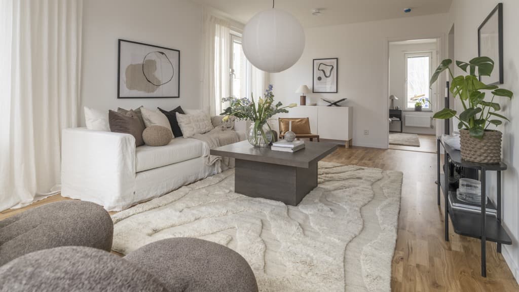 Ett vardagsrum med matta, soffa, soffbord och sittpuffar i vita, grå och beige toner. Gröna växter och parkettgolv.