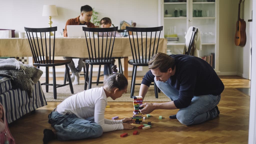 En bonuspappa spelar ett spel med klossar med en bonusdotter på golvet i ett vardagsrum. Flickans syster och pappa sitter vid ett bord i bakgrunden.