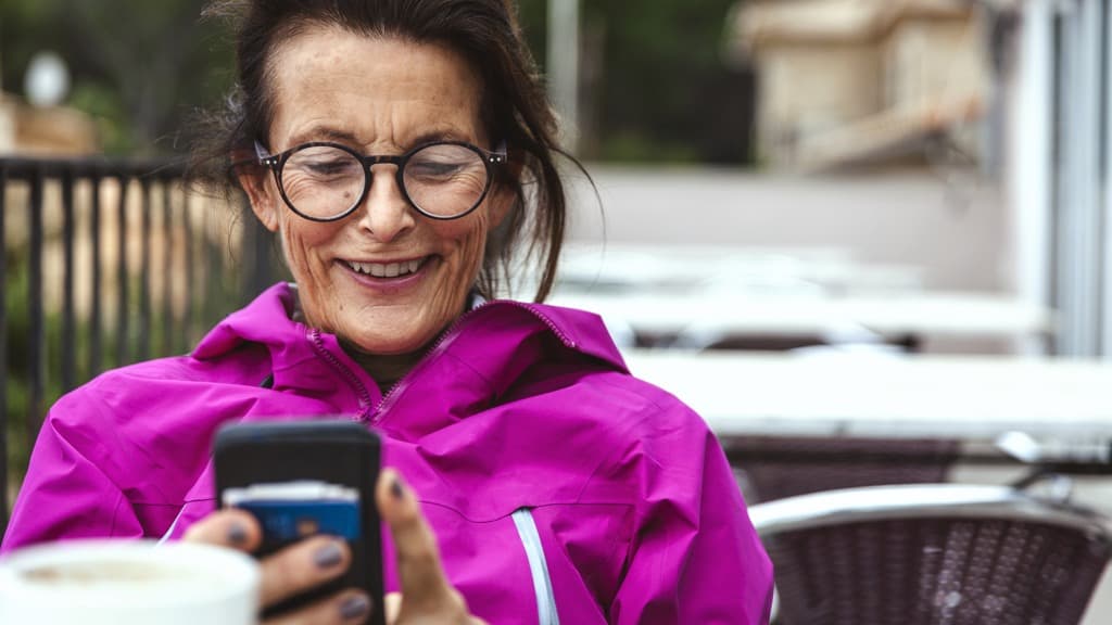 En äldre kvinna sitter vid ett bord på ett kafé med en kopp kaffe och en mobiltelefon.