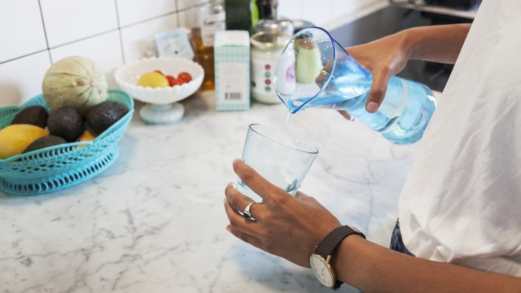 Bruna kvinnohänder häller vatten i ett glas från en karaff vid en köksbänk