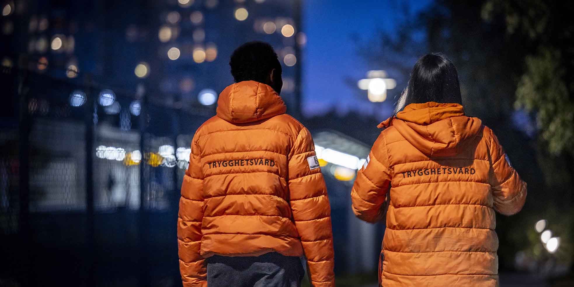 Två trygghetsvärdar i orange jackor går på gatan i mörk kvällsmiljö med hus i bakgrunden.