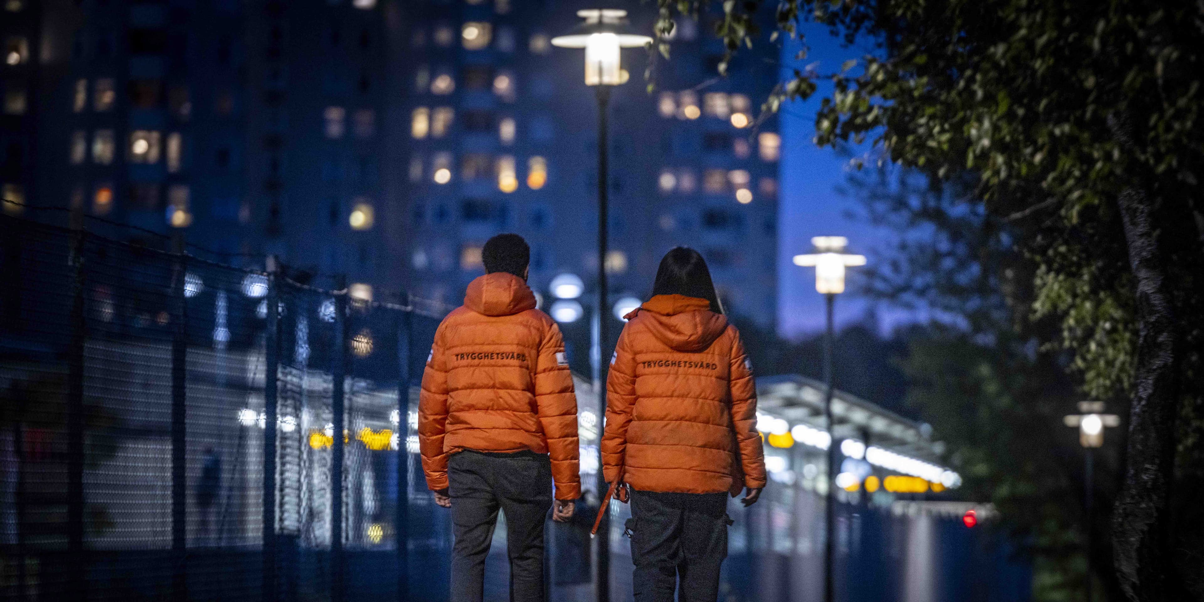 Trygghetsvärdar i Farsta med orange jackor går i mörk miljö med hus med fönster där ljus strålar ut.