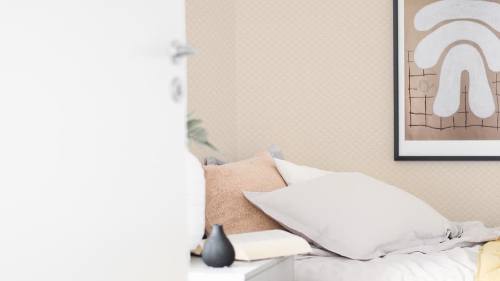 En öppen dörr mot ett sovrum med blekrosa tapet och en säng med kuddar i beige och blekrosa.