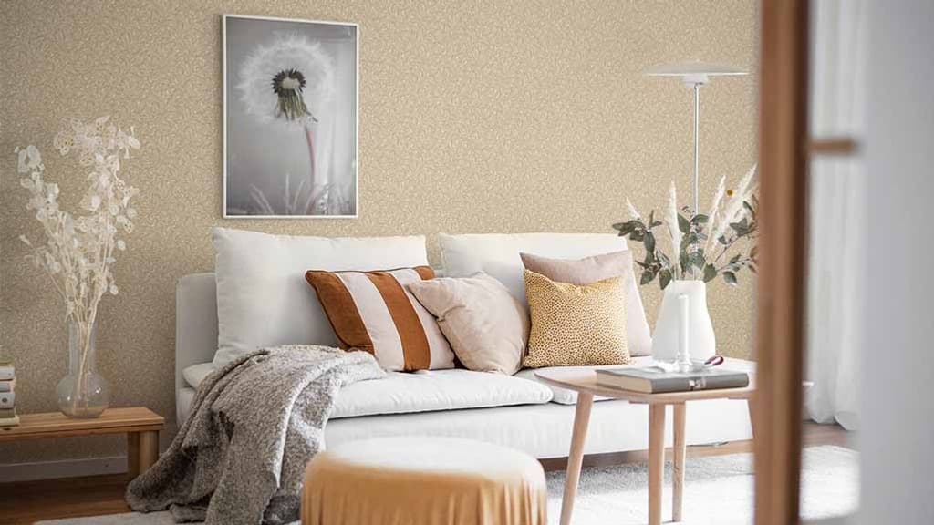 Ett vardagsrum med småmönstrad tapet i en persikobeige ton. En svartvit tavla med maskros boll på väggen, en vit soffa med kuddar i beige, blerosa och orangebruna toner. Sitt puff vid vardagsrumsbordet med bok och vas med torkade gräsvippor och gröna kvistar.