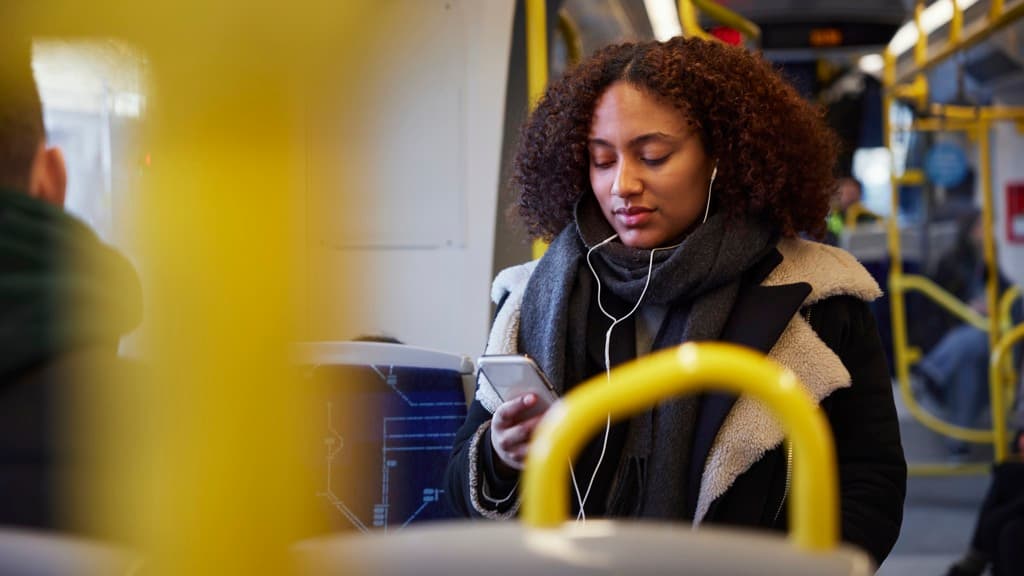 En ung kvinna i kollektivtrafiken som har hörlurar och tittar på sin mobiltelefon.