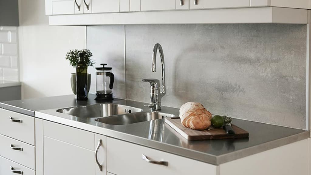 Stänkskiva av kompaktpanel som ser ut som grå cement över bänkskiva i rostfritt utseende. En skärbräda med bröd, kaffepress, olja och en liten växt på köksbänken.