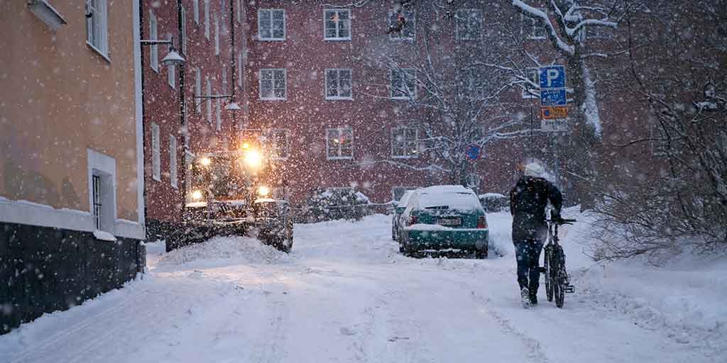En snöig bostadsgata med liten plogmaskin på trottoaren, parkerade snöiga bilar och en cyklist.