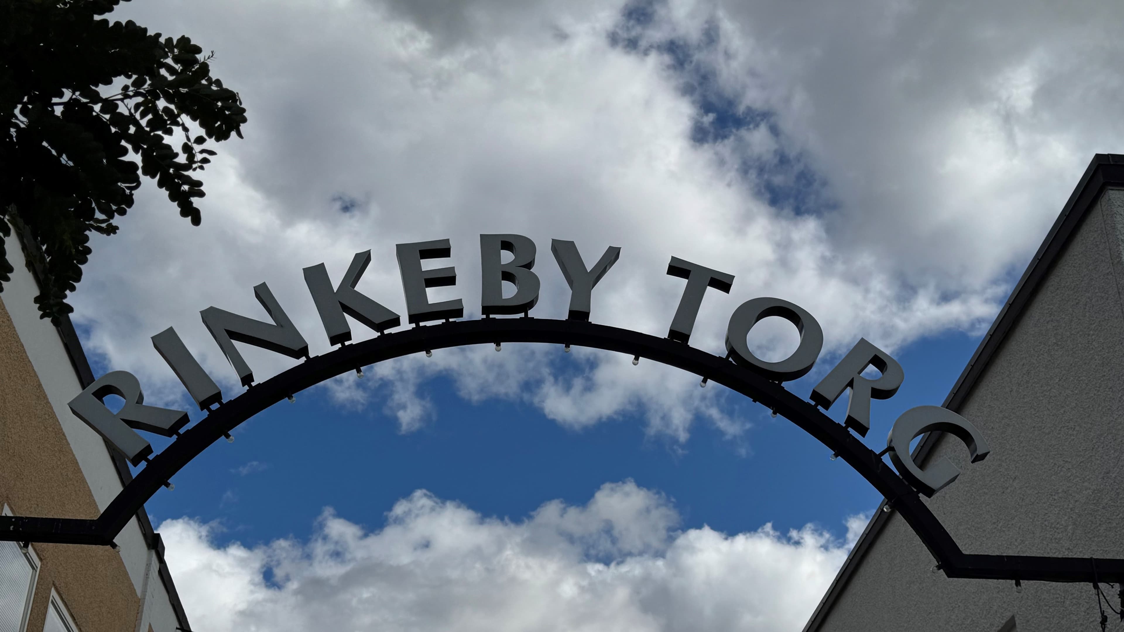 En skylt med texten "Rinkeby torg" mot blå himmel med vita moln