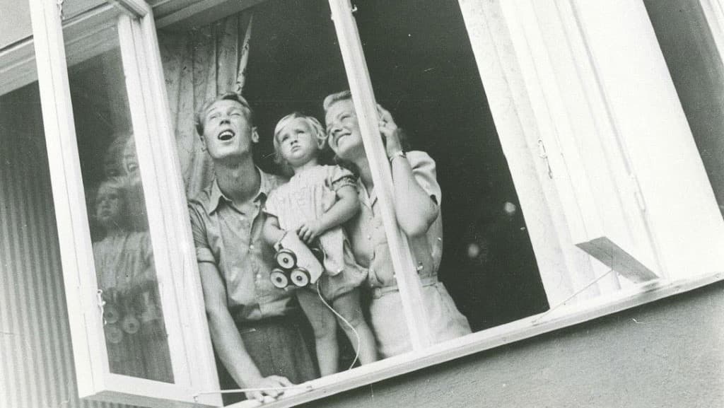 En familj tittar ut från ett fönster i ett flerfamiljshus på 50-talet.