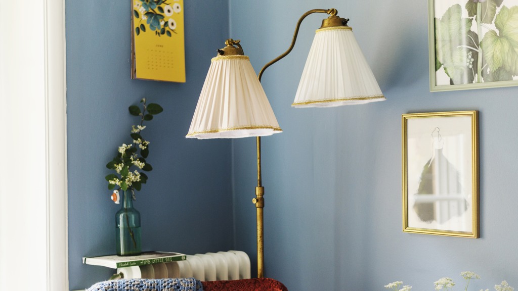 Ett rumshörn med duvblå väggar, tavlor - bland annat en gul tavla med blomstermotiv. En turkos flaska med blommande kvist och en tvåarmad retrolampa syns också i bild.