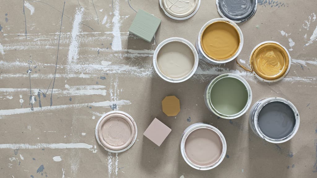 Öppnade färgburkar i blekrosa, senapsgul, lindblomsgrönt och beige på golvpapp med vita målarstreck på.